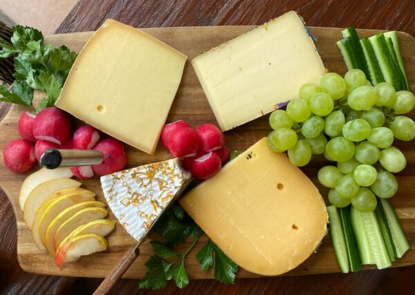 image of a variety of cheese with veggies and fruit on a cutting board.