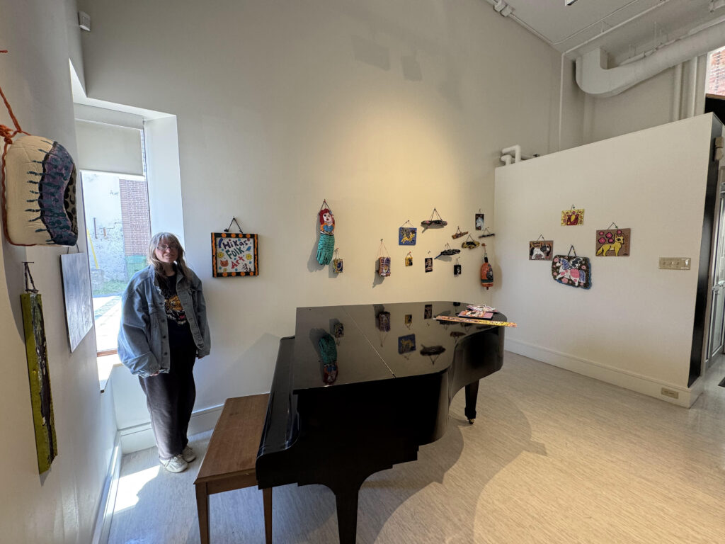 image of woman with long light hair standing next to folk art displayed on a gallery wall.
