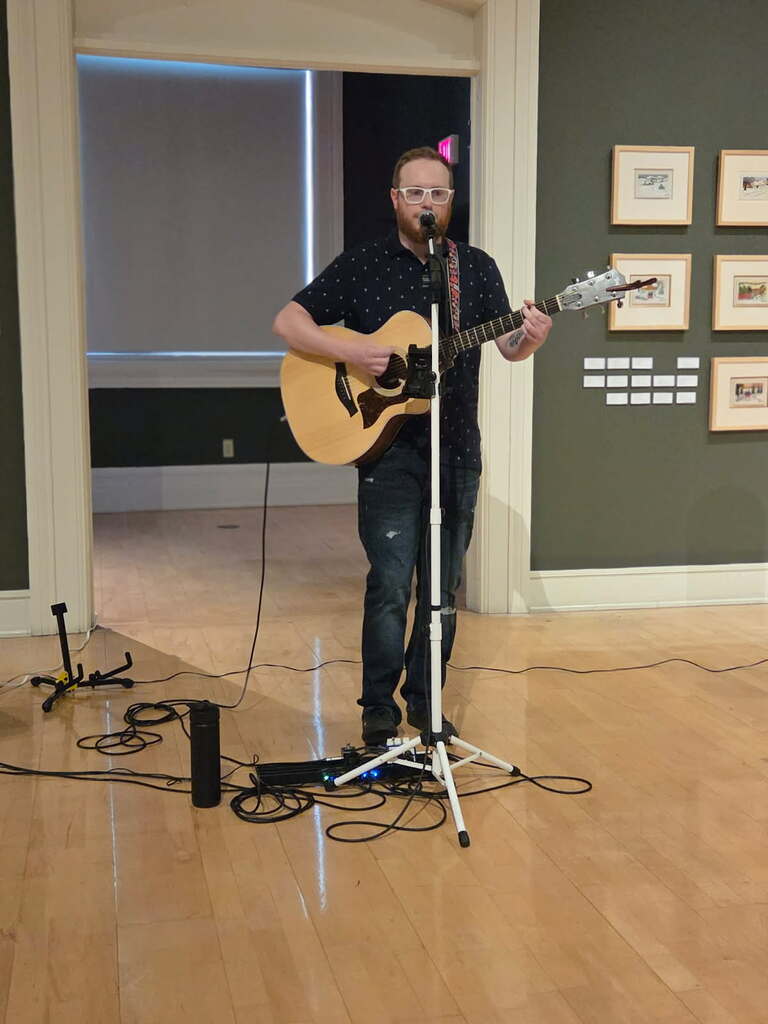image of a man with red hair and a beard, white glasses, dark short-sleeved shirt, playing a guitar.